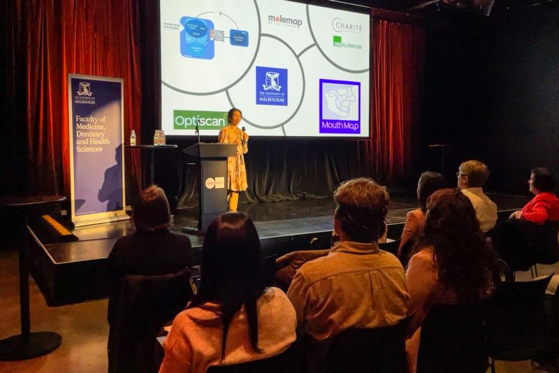 Dr Tami Yap speaking at an event for Melbourne Connect Innovation Week. Behind her is a project screen with the partners she collaborated with on her project, MouthMap.