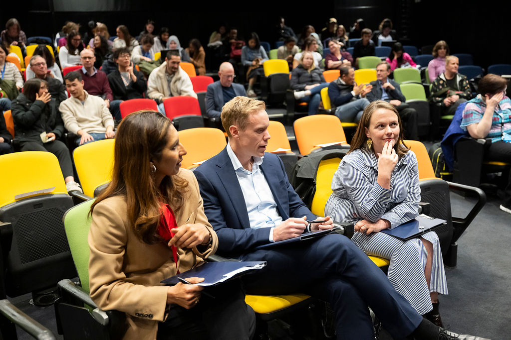 Two women and a man sit on chairs talking to each other. A crowd of people sit behind them.
