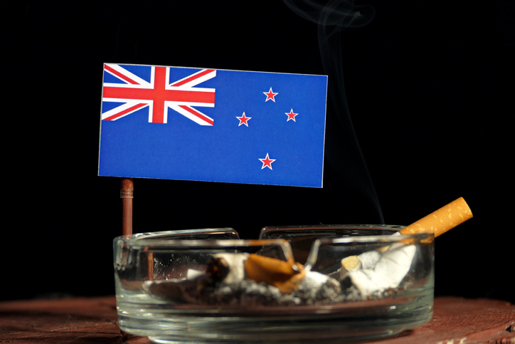 A small New Zealand flag is placed above an ashtray with a discarded cigarette on a small wooden table. There is a black background.
