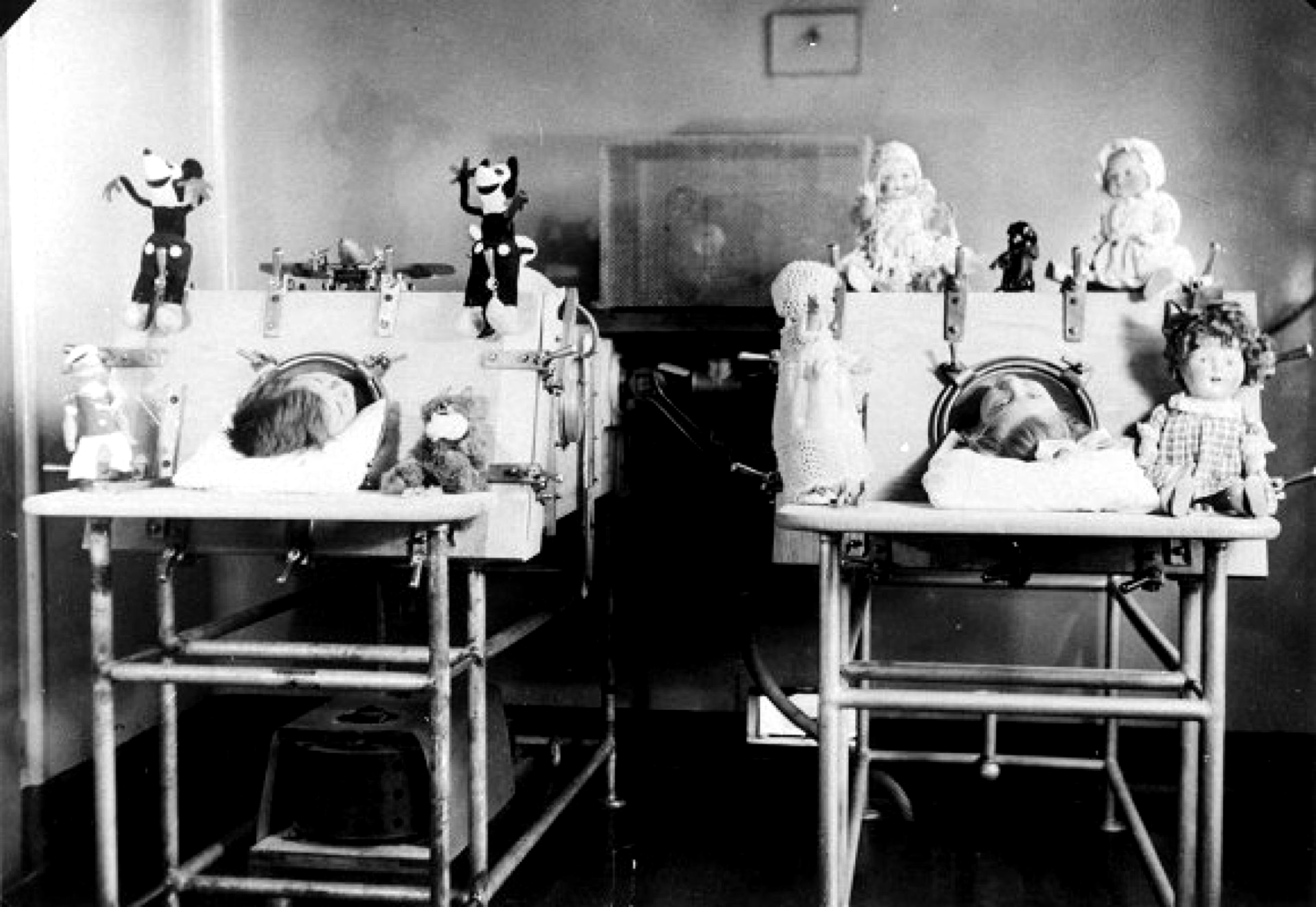 Two young polio patients lie in their beds with their torsos encased in in iron lungs, their heads surrounded by toys they are unable to play with.