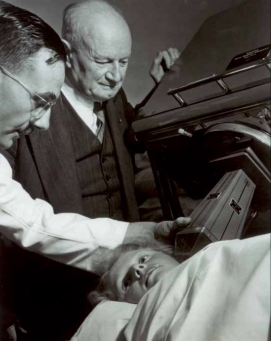 Sir Peter MacCallum and a colleague standing next to a young patient lying on a linear accelerator, c. 1959