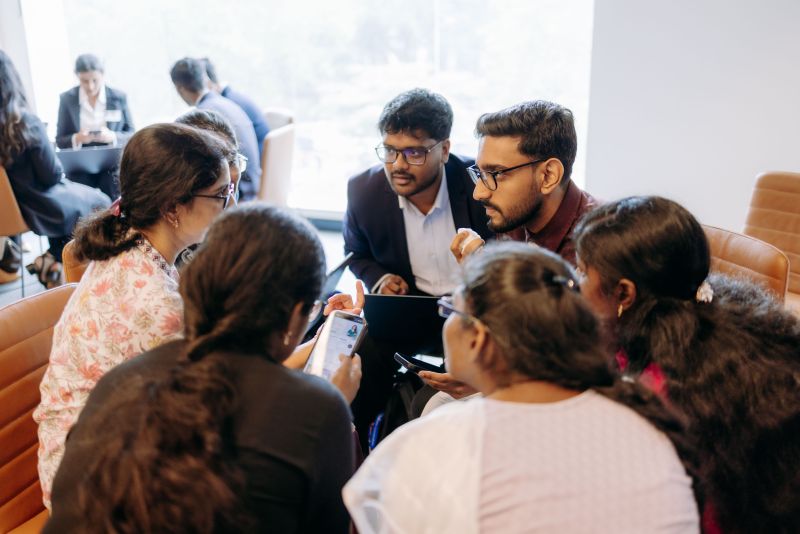A team of students sitting in a circle discuss their case. They appear focused and determined. Some of the students are holding mobile phones, and one has a laptop in his lap.