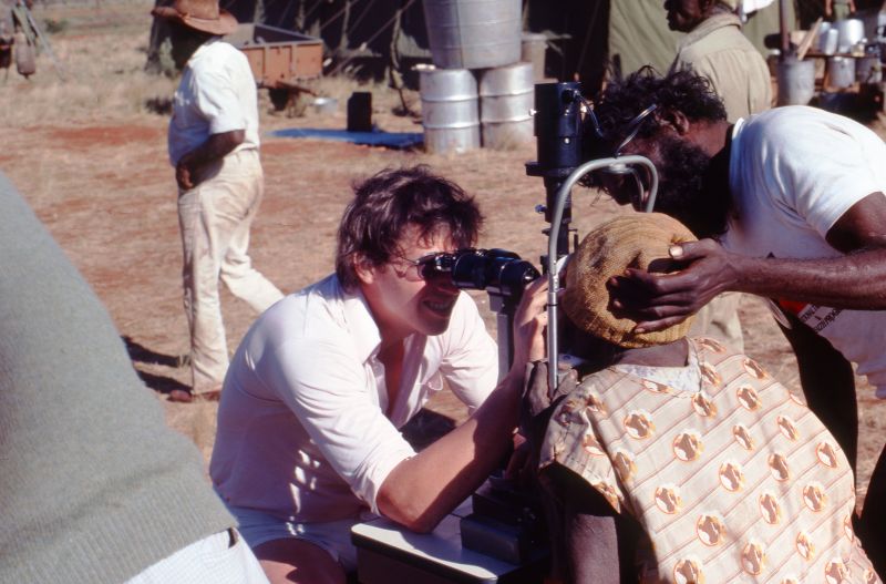 Professor Hugh Taylor using a magnifying device to inspect the eye of a patient. The inspection is happening outdoors, with dry grass and some vehicles and barrels in the background. The photo is taken on a film camera, featuring a young Professor Taylor. A man stands next to the patient, placing his hand on the back of her head and supporting her during the inspection. 
