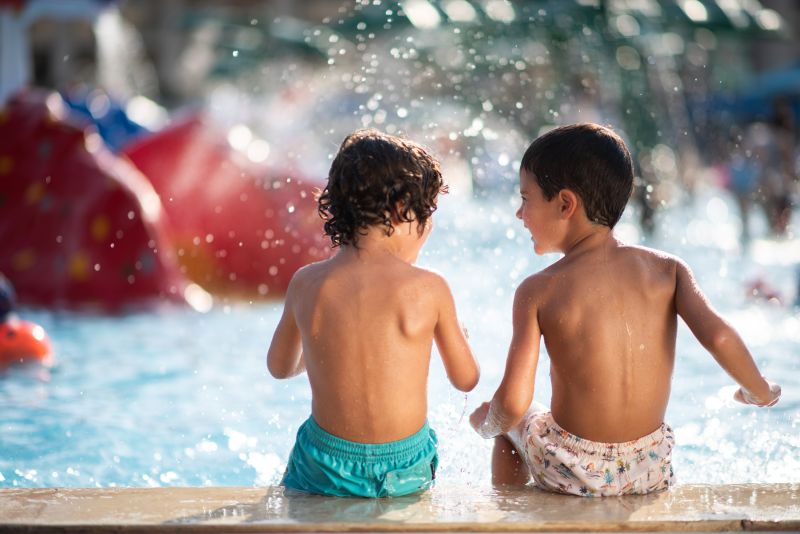 Two young children sit side by side at the edge of a shallow pool, splashing water and playing in a sunny outdoor water park.