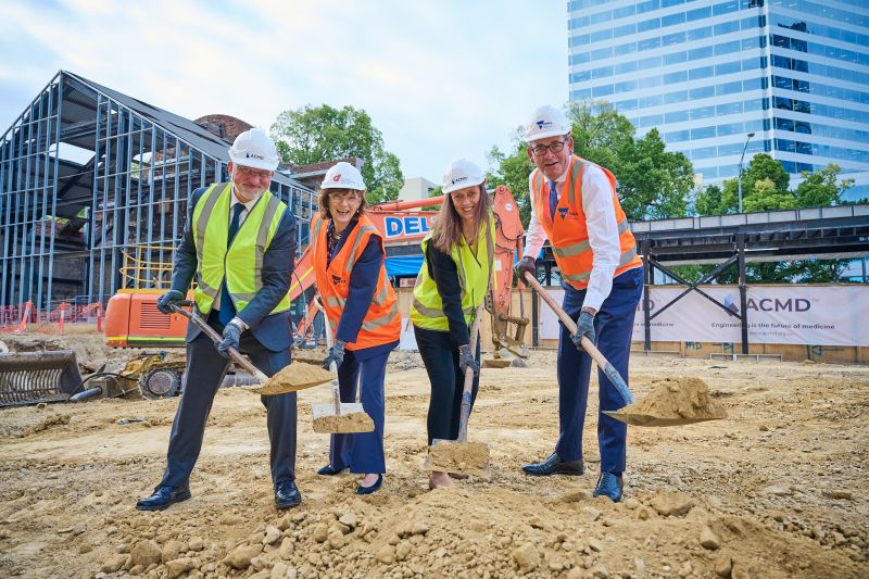 Dr Erol Harvey, Minister Mary-Anne Thomas, Nicole Tweddle and Premier Dan Andrews at the site of the new ACMD facility. They are each holding a shovel with dirt. The background is a construction site, with dirt and gravel on the ground, and some minor scaffolding.