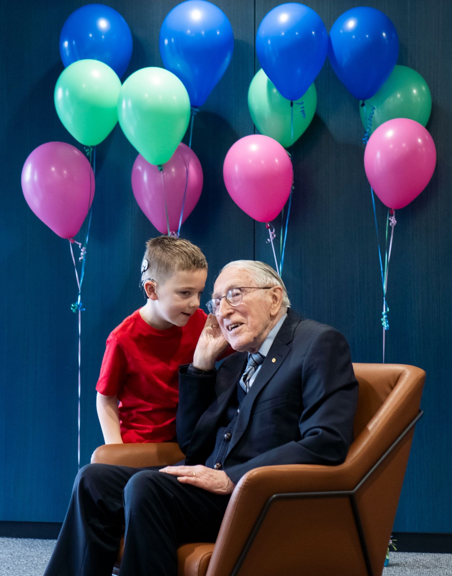 Professor Graeme Clark AC sits in front of some balloons with six-year-old cochlear implant recipient Hudson. Hudson is whispering something in Professor Clark's ear.