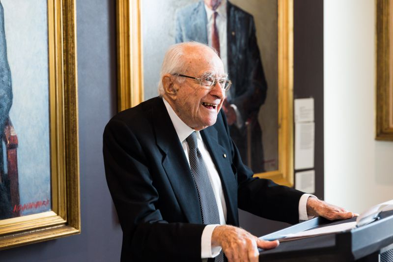 Emeritus Professor Derek Denton speaking at a podium. In the background there are two painted portraits of distinguished academics with golden frames. Emeritus Professor Denton is wearing a black suit with a spotted tie. It is a recent photo. 