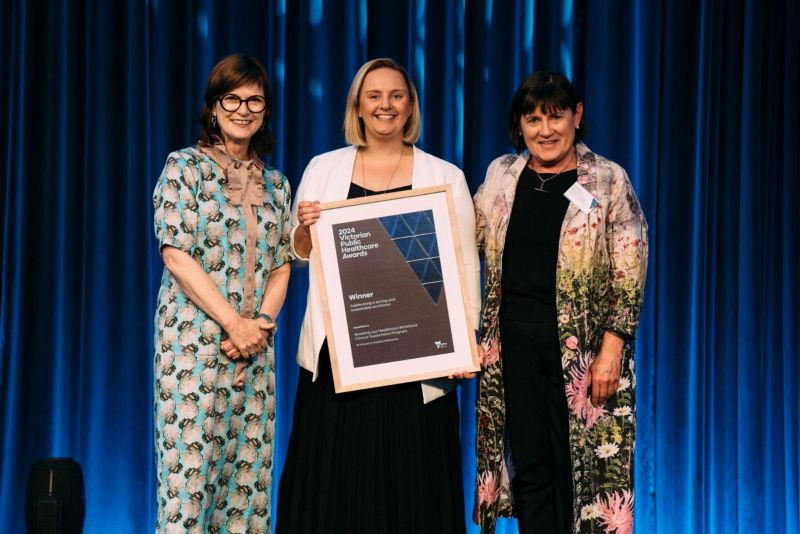 Mary-Anne Thomas, Victorian Minister for Health, with Jade Murphy and Fiona McKinnon at the awards ceremony. Jade is holding the award, which is a framed certificate. There is a blue lit curtain in the background.