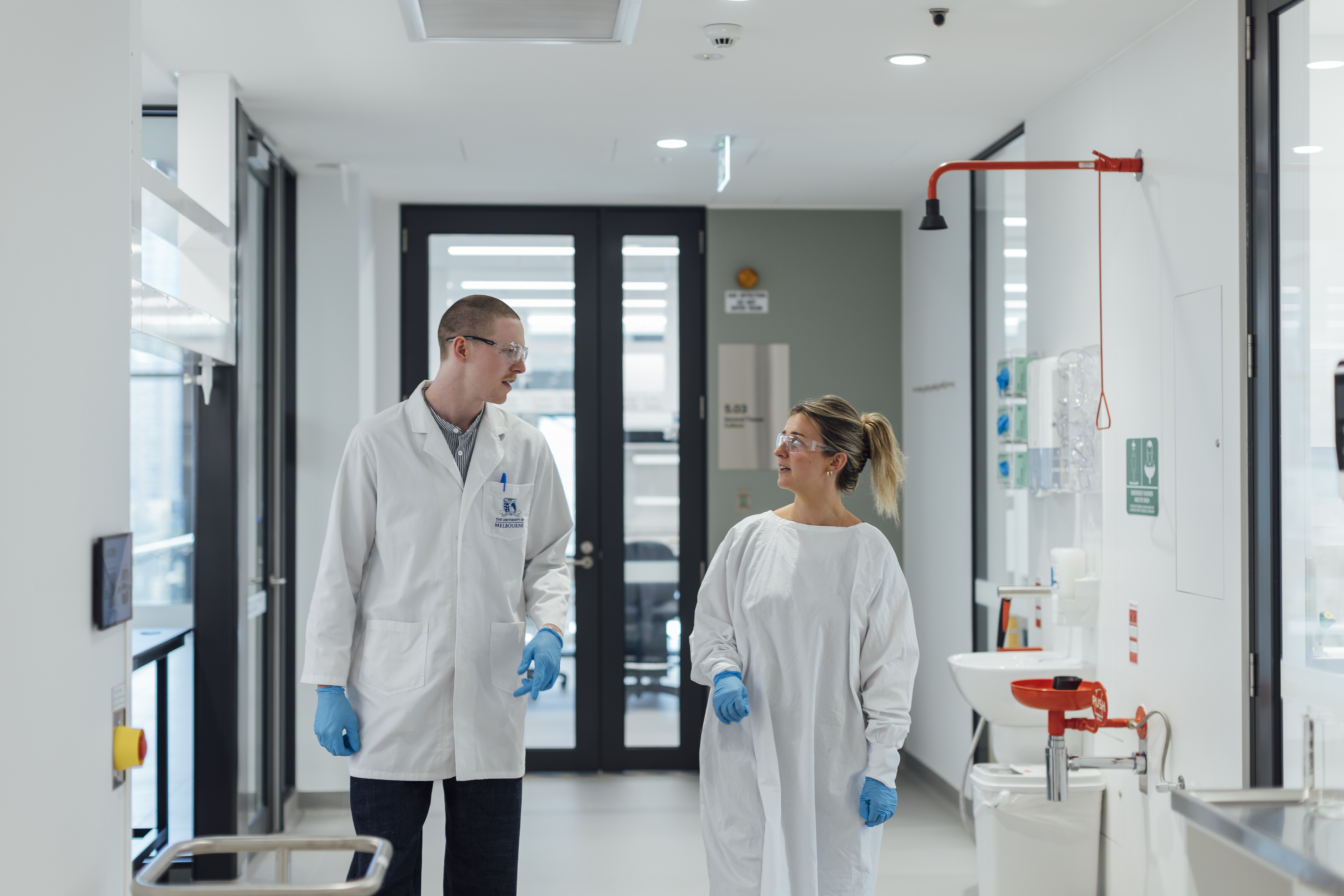 PhD student William Humble walks alongside a female colleague down the hallways of ACMD. They are both wearing lab coats and protective goggles.