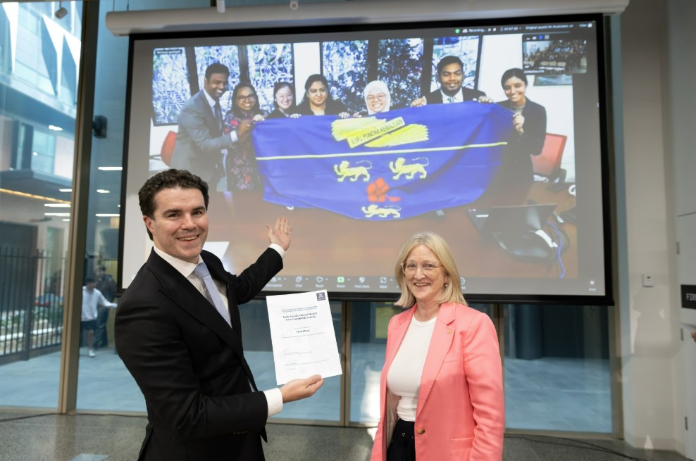Tim Watts and Jane Gunn holding a certificate in front of a projector screen, which shows the winning team from Universiti Malaya via Zoom.