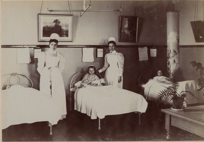 Two female nurses stand looking at the camera while three young patients lay in beds in the Summer Ward of the Children's Hospital, 1895.