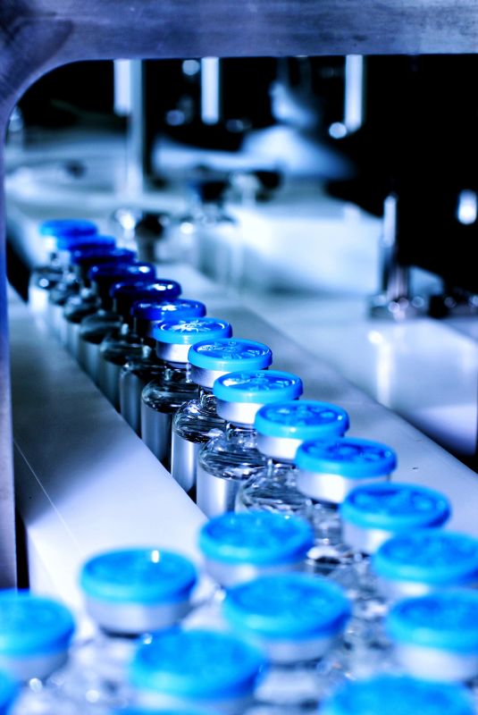 A row of vaccine vials moves along a production line in an industrial laboratory setting.