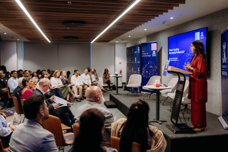 The Hon Lisa Singh standing at a lectern on a slightly raised stage. She is addressing a seated audience of about 40 people. The room is modern and grey, with a display screen and university-branded banners on stage.