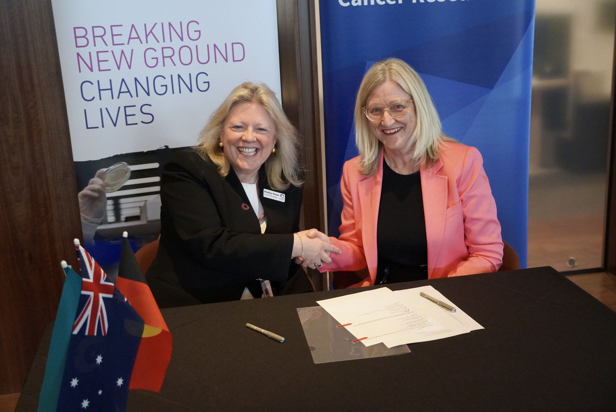 Professor Shelley Dolan, Chief Executive of the Peter MacCallum Cancer Centre, shakes hands with Professor Jane Gunn AO, Dean of the Faculty of Medicine, Dentistry and Health Sciences, at the signing of the MoU. Shelley and Jane are seated at small table, on which the MoU documents are placed. 