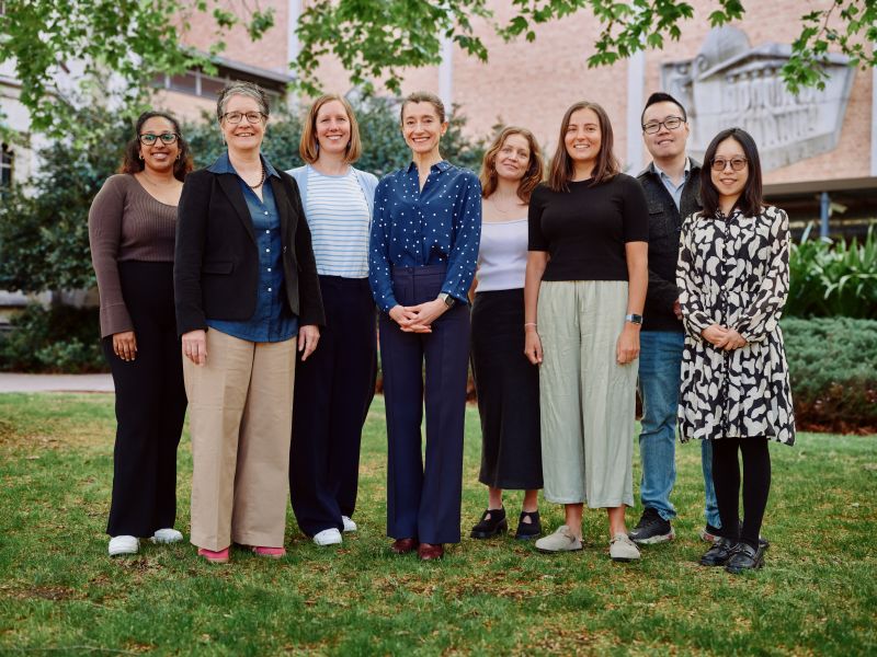 The Melbourne Research Group standing on a lawn, with a brick building in the background. There are 7 team members altogether.