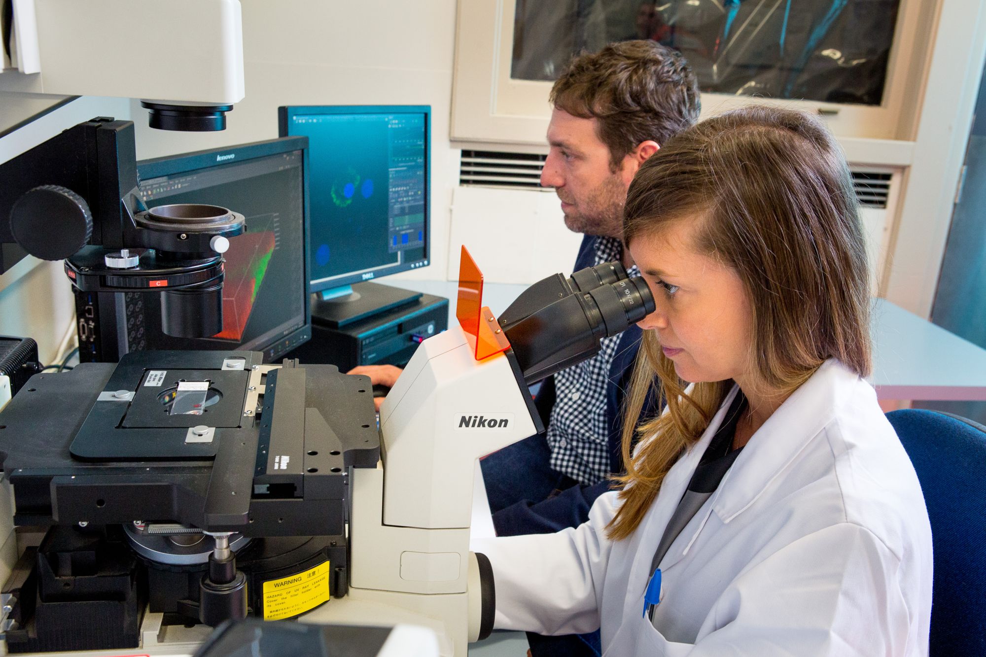 Associate Professor Serena Duchi looks through a microscope in the lab while a colleague works beside her at a computer monitor.