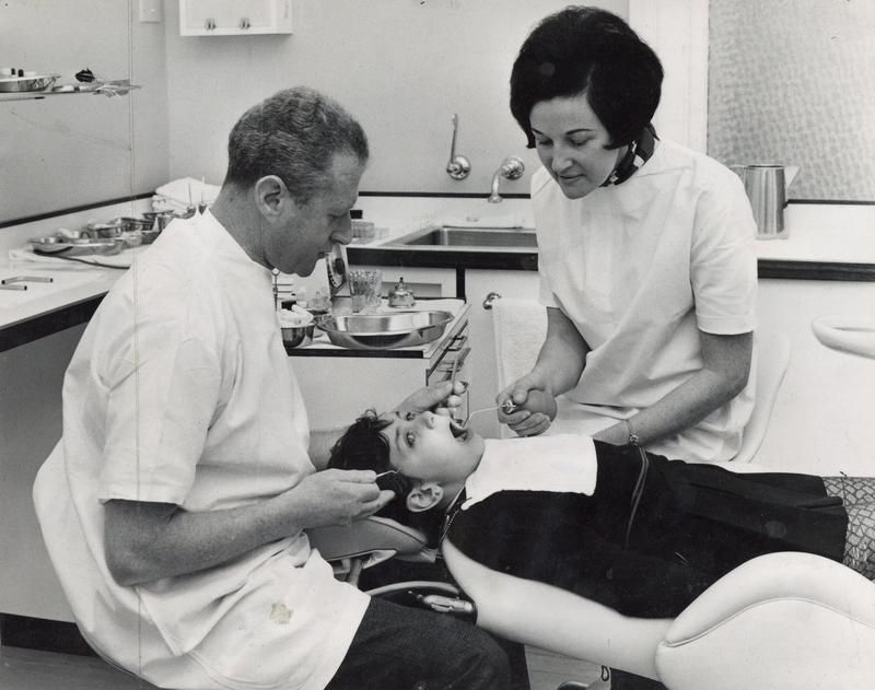 Photograph of a dentist examining a young child's teeth with a dental nurse assisting.